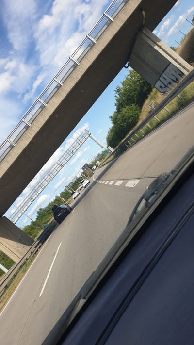 The image is taken from a car driving on a highway with an overpass visible in the foreground. The sky is blue with white clouds. The highway has multiple lanes with cars and trucks driving on it. The overpass is made of concrete and has a metal railing. There are trees and bushes on the side of the highway. The image is slightly tilted. The specific location in the city of Hodkovice, Czechia is not provided in the image.