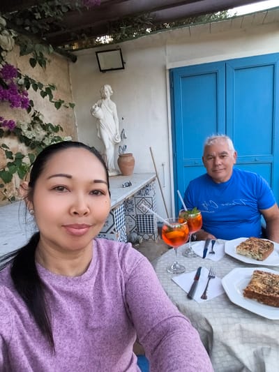 A medium eye-level indoor shot shows a light-skinned Asian woman and a light-skinned older man at a table with food and drinks in San Ġwann, Malta. The woman is in the foreground, facing toward the camera. She is wearing a long-sleeved lavender shirt, and her black hair is pulled back. Behind her and to the right is the man, wearing a blue short-sleeved shirt. They both are smiling. In front of them on the table are two plates of what appears to be bread and two glasses of orange liquid with straws. Each place setting has a fork, knife, and white napkin. Behind the man is a double blue door with a white wall. To the left of him is a white statue of a woman holding an urn, with purple and white flowers and vines growing up the wall. Above the statue and extending to the right is a roof with slats.
