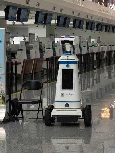 A white and blue robot with a touchscreen display and cameras is positioned in the foreground of an airport or train station check-in area. The robot has "ARTROBOT" and Chinese characters printed on its side. It is standing on a tiled floor with reflective surfaces, and a black folding chair is to its left. In the background, rows of check-in counters with electronic displays and security cameras are visible, along with signage indicating check-in zones labeled E04 through E15. The environment appears to be an indoor public transportation hub, likely in Guangyang District, China, given the presence of Chinese text on the robot.