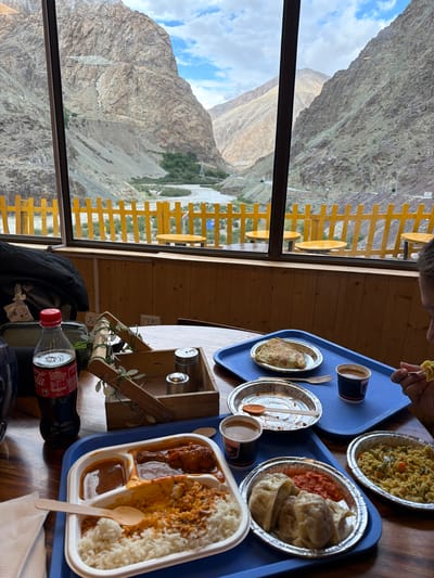 The image captures a meal enjoyed at a restaurant with a scenic view of the mountains and a river in Kiari, India. The foreground features a wooden table laden with food on blue trays. On the left, a bottle of Coca-Cola is placed next to a small wooden basket holding salt and pepper shakers. A white meal tray contains rice, curry, and a chicken dish. Adjacent to it, another silver plate holds steamed dumplings (momos) with a red sauce. To the right, a portion of noodles with vegetables is served in a silver oval dish. In the background, a large window reveals a breathtaking view of rugged mountains under a cloudy sky, with a river flowing through the valley. Yellow fencing and outdoor seating are visible beyond the window. A person's hands are visible in the upper right corner, holding a fork and eating from a plate.