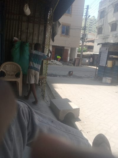 The image displays a sunlit urban street scene in Tudor ward, Kenya, viewed from a shaded, ground-level perspective. In the immediate bottom-left foreground, a blurred portion of a human leg, clad in light grey trousers, is visible. To the left, a white plastic chair is positioned next to two large green fabric sacks leaning against a corrugated metal wall. A white cap hangs from this wall. A young male, facing away from the camera, stands holding a metal gate or barrier with both hands. He wears a horizontal striped t-shirt and light-colored shorts, with bare feet.
The midground features a light-colored, brick-paved street, illuminated by direct sunlight. A concrete block element rests on the left edge of the pavement, partially shadowed. A brown and black chicken with a red comb is present on the street surface, oriented towards the right. Further down the street on the left, a building under construction or renovation displays an open doorway with dark brown panels; a person in a high-visibility yellow-green vest is partially visible inside, near a pile of white material. On the opposite side of the street, a structure resembling a stall displays a white sign with black text and a yellow container. An individual is faintly visible near this stall. A utility pole laden with multiple black cables stands on the right.
The background reveals several multi-story buildings under a clear sky. A prominent beige-colored building with blue-framed windows exhibits visible surface irregularities, indicating unfinished construction or age. Distant taller buildings are also discernable beyond the immediate structures. The street surface shows scattered dirt and debris.