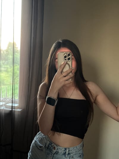 A young woman with long, dark hair takes a selfie in a mirror. She is wearing a black strapless top and ripped denim shorts. On her left wrist, she has a silver smartwatch with a black screen. She is also wearing a silver ring on her left ring finger and a delicate silver necklace. The background shows a window with sheer dark curtains and a view of greenery outside. The room appears to be dimly lit, with natural light streaming in from the window. This image was captured in Horenychi, Ukraine.