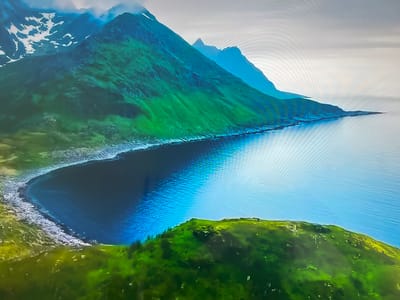 The image shows a coastal landscape with mountains. The mountains are green with some snow on the higher peaks. A body of water, likely the sea, extends into a bay. The water is blue with reflections. The coastline is rocky. There is green vegetation in the foreground. The sky is overcast.
The location in the city of Basingstoke and Deane, United Kingdom is not correct for this image. The terrain is mountainous. The city of Basingstoke and Deane has no coastline or mountainous regions.