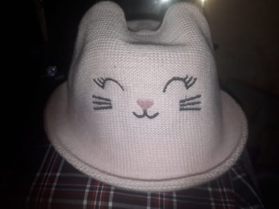 A light pink knitted hat with cat-like ears and embroidered facial features is centered in the frame, resting on a red and black plaid fabric. The hat features two embroidered eyelashes on each side of a pink embroidered nose and a black embroidered mouth. The background is dark and indistinct, suggesting an indoor setting. The location in Zilupe, Latvia, is not discernible from the image.