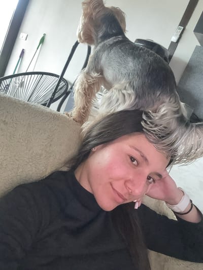A woman lies on a beige-colored sofa, wearing a black long-sleeved shirt. A small Yorkshire Terrier-type dog stands near her head, partially on the sofa and perhaps partially on her head. Her right hand is raised, seemingly touching the dog. A black metal chair with a circular, webbed back is visible behind the dog. In the background, cleaning tools like mops or brooms lean against the wall. The image might be taken indoors, possibly a residence. No specific landmark is discernable to ascertain the location as Lipnik, Bulgaria.