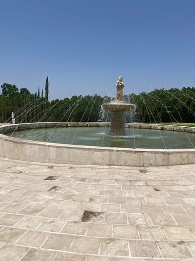 The image shows a fountain in Tehuixtla, Mexico. The fountain consists of a multi-tiered basin with a statue on top, surrounded by jets of water spraying outwards. The fountain is positioned next to a paved area made of light-colored rectangular stones. In the background, dense green trees stand under a clear blue sky.