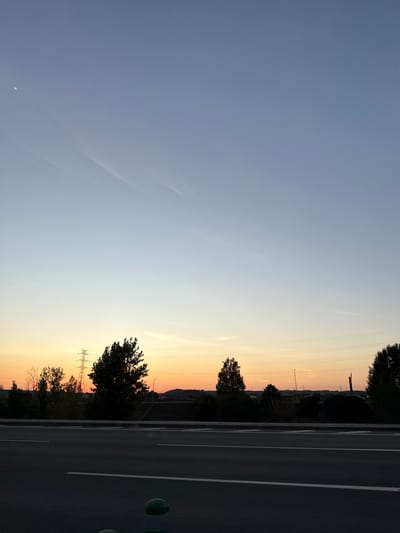 This media file captures a tranquil landscape scene, most likely during either sunrise or sunset, with the location context being Santarém, Portugal. The image is dominated by a vast, clear sky that presents a beautiful color gradient. It transitions from a soft blue-grey at the top to warm shades of orange and yellow closer to the horizon, indicative of dawn or dusk. Faint, wispy clouds or contrails are subtly visible in the upper portions of the sky, and a very small, crescent-like white speck, possibly the moon, can be seen in the upper left corner.
Along the right side of the frame, a dark asphalt road stretches into the distance, marked with clear white lines that delineate lanes or shoulders. Several green, circular objects, possibly bollards or road markers, are spaced along the immediate edge of the road.
The horizon is defined by the dark silhouettes of numerous trees and a distant, undulating landscape, which could be hills or the outline of a distant town. Overlaid against the vibrant sky, slender power lines and utility poles are visible, hinting at human infrastructure blending into the natural environment. No people or visible text are present in the image. The overall atmosphere conveyed is one of peace and natural beauty under a transitioning sky.