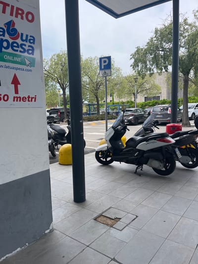 The image captures a scene in Castiglione delle Stiviere, Italy, featuring a paved area with several parked scooters and cars. In the foreground, a white and black scooter is prominently displayed, with another scooter partially visible to its right. To the left, a white pillar with red and blue lettering that reads "TIRO a tua spesa" and "ONLINE" is present, along with a red arrow pointing upwards and the text "50 metri". Further back, a yellow cylindrical object, possibly a bollard, is situated near a dark-colored scooter. A blue and white parking sign with a "P" and a bicycle symbol is visible in the background. Several vehicles, including a dark SUV, a grey hatchback, and a white van, are parked in the distance. The environment appears to be an outdoor public space, possibly a parking lot or commercial area, with trees and buildings visible behind the parked vehicles. The sky is overcast, suggesting a cloudy day.