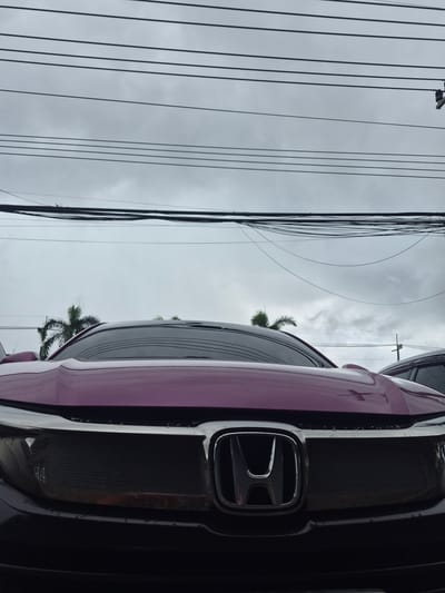 A low-angle shot captures the front of a purple Honda Civic car against a cloudy sky in Ban Yid, Thailand. The car's hood and grille are prominent, with the Honda logo clearly visible. Overhead, a dense network of power lines crisscrosses the frame, silhouetted against the overcast sky. Palm trees can be seen in the background, suggesting a tropical or subtropical environment. The overall atmosphere is somewhat muted due to the cloudy weather. No people are visible in the image.