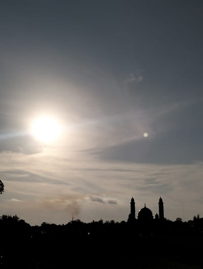 The image depicts a panoramic view of a silhouetted landscape under a bright, partly cloudy sky, likely at sunrise or sunset, in what is identified as Keudetambue, Indonesia. The dominant feature in the upper-left quadrant is an intensely bright, overexposed sun, casting strong lens flare effects and illuminating scattered clouds across the sky, which transitions from pale yellow-white near the sun to a darker grey-blue at the top right. A smaller, fainter circular light anomaly is present in the mid-right sky. Several small, dark specks, consistent with birds in flight, are visible in the upper-left near the sun.

The lower third of the scene is rendered in complete silhouette, showing a horizon line of dense vegetation. Prominently centered within this silhouette is a mosque, distinguishable by its large central dome and two tall, slender minarets positioned symmetrically on either side. To the left of the mosque's silhouette, a plume of dark, vertical smoke rises from behind the treeline before dissipating horizontally into the sky, intermingling with clouds. The immediate foreground below the silhouetted trees and mosque is an undifferentiated dark expanse, possibly a field or open ground. No individuals are discernible.