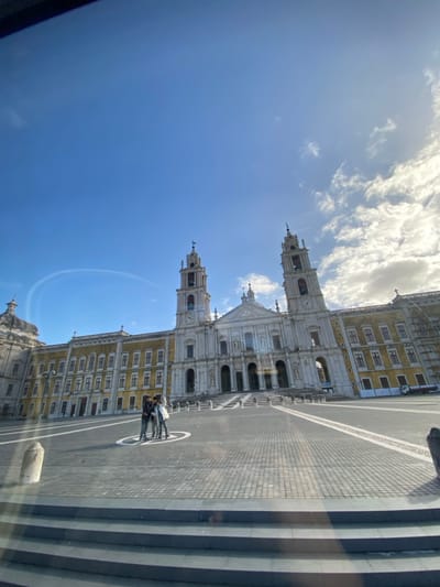 The image depicts the Mafra National Palace in Mafra, Portugal, under a clear blue sky with scattered white clouds, predominantly on the right. The sun is bright, casting shadows across the foreground.
The central subject is the grand Baroque complex of the Mafra National Palace, featuring a prominent white stone basilica with two symmetrical bell towers. Each tower is topped with a weather vane and displays a clock face. The basilica's facade includes multiple columns, arches, and sculptural elements, with a wide set of steps leading up to its main entrance. Flanking the central basilica are two extensive wings of multi-story buildings, characterized by their yellow-ochre facades and numerous windows with gray detailing. Scaffolding is visible on the far right section of the palace's right wing, indicating ongoing work.
In the large, paved square (Terreiro D. João V) in front of the palace, three individuals are standing together near the center. They are positioned within a circular marking on the dark grey cobblestone-like pavement, which features lighter contrasting lines forming a geometric pattern. The individuals are casually dressed. A small white utility vehicle is parked near the base of the palace's right wing.
The foreground shows several dark, horizontal steps, suggesting the image was captured from a slightly elevated position, possibly through glass, as faint reflections are visible across the lower portion of the frame. A subtle, curved white contrail is visible in the upper left section of the blue sky.