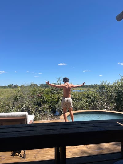 A shirtless man in light-colored shorts stands on a wooden deck with arms outstretched, appearing to look out over a marshy landscape in what is likely the Khwai region of Botswana, as the foliage suggests a wetland ecosystem. A circular wooden pool is located to the right of the man on the deck. A wooden structure frames the right side of the image, partially obscuring a glimpse of a light-colored couch. The sky is clear blue with a few scattered clouds. The scene is brightly lit, suggesting a sunny day.