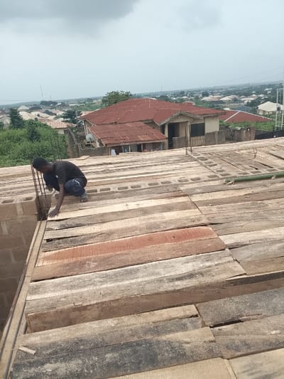 The image depicts a construction site in the city of Arun, Nigeria, under an overcast sky. In the foreground, a male individual wearing a black t-shirt, dark trousers with light-colored lettering on the left thigh, and light-colored footwear, crouches on a wooden plank formwork. He is positioned next to a concrete block wall with vertical steel rebar protruding upwards. His left hand rests on the wooden planks, while his right hand is near the rebar. The construction platform is composed of wooden planks laid over pre-cast concrete blocks, with exposed horizontal rebar sections. A green bamboo pole is visible on the right side of the planks. The background showcases a dense residential area, characterized by numerous buildings, many with red corrugated metal roofs, interspersed with green trees and vegetation. The cityscape extends to a hazy horizon, with distant structures faintly discernible.