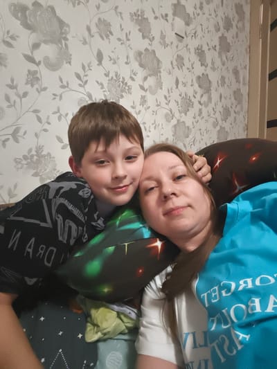 This is a selfie taken by two people, a boy and a woman, likely mother and son, lying down and posing for the camera. The boy is in the foreground, leaning his head on a pillow, with his arm resting on the woman's shoulder. The woman is behind him, resting her head on another pillow and looking at the camera with a slight smile. Both are casually dressed. The boy wears a black t-shirt with a graphic print in grey. The woman is wearing a bright blue t-shirt over a white t-shirt.
They are in an indoor setting, likely a living room or bedroom. The background features floral wallpaper with a light, neutral color. There are several pillows around them, including one with a dark background and glowing red and yellow star-like patterns, and another with a dark background and a green galaxy-like print. A folded light green blanket is visible on the lower right.
The lighting suggests it is daytime, possibly in the afternoon, as the light is soft and diffused. The overall mood appears relaxed and affectionate, with both subjects smiling or appearing content.
Visible text includes the words "И АЯО" and a series of letters on the boy's shirt, and "FORGET ABOUT JUST LIVE" written in reverse on the woman's blue t-shirt. The setting is identified as Malaya Valyaevka, Russia.