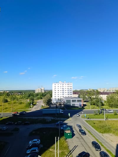 The image depicts a daytime aerial view of a street intersection and surrounding buildings in Kokhma, Russia. The sky is clear and blue with a few scattered clouds. A multi-story white apartment building with numerous windows is prominently featured on the right side of the image. Adjacent to it is a modern, low-rise commercial building with a grey facade and glass windows.
A road runs through the scene, with several cars parked on its shoulders and driving on it. A large blue semi-trailer truck is parked on the right side of the road. On the left side of the road, a green dumpster is visible. The area is characterized by a mix of paved surfaces, green grass, and trees. In the distance, more buildings and a treeline are visible, suggesting a suburban or semi-urban environment. There are street signs and traffic signals present at the intersection. No people are clearly identifiable in the image. The overall scene suggests a typical day in a Russian city.
