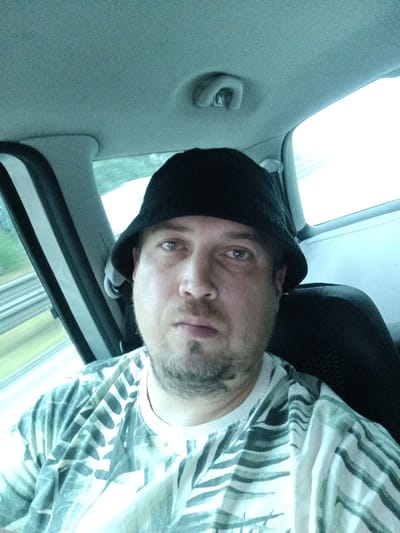 This is a first-person, close-up selfie of a man wearing a black bucket hat and a light-colored t-shirt with a dark, abstract pattern. The man has light skin, stubble, and is looking directly at the camera with a neutral expression. The background shows the interior of a car, with a window on the left revealing a blurred green and white landscape suggesting movement. The ceiling of the car is visible at the top of the image. The lighting is bright, and the overall mood is casual, possibly indicating the man is a passenger on a journey. The image has a slight blueish tint, which may be due to the lighting conditions or the camera used. There is no visible text in the image. Given the location context of Ostředek, Czechia, it is plausible the man is traveling within or through this region. The weather appears to be overcast or the time of day is such that the natural light is diffused.
