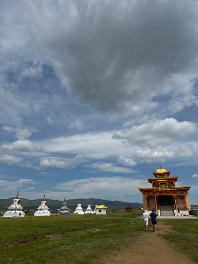 In Orongoi, Russia, a sunny day with scattered clouds frames a Buddhist complex. A golden-roofed, red and yellow temple stands on the right, its entrance accessed by a set of stairs. Two men, one in a white hooded sweatshirt and shorts, the other in a dark t-shirt and shorts, walk away from the camera towards the temple. In the middle ground, a line of white stupas, some adorned with gold and blue accents, stretches across a grassy field. A smaller white building with a yellow roof is visible to the left of the temple, and distant mountains form the backdrop under a dynamic sky.