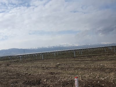 Rows of solar panels stretch across a barren, brown landscape towards snow-capped mountains under a cloudy sky. Support structures hold the panels aloft. A marked post stands in the foreground. A transmission tower is also visible in the background.