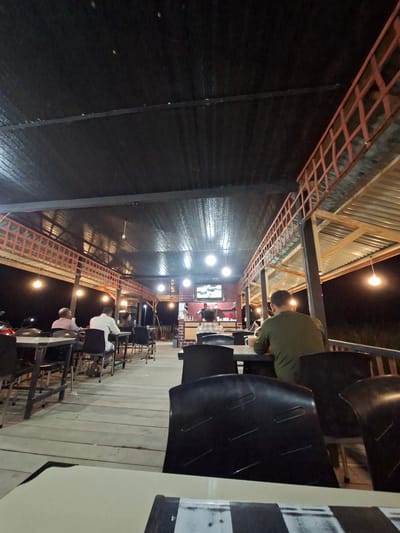 The image depicts an outdoor seating area at night, possibly a restaurant or cafe, located in Puuk, Indonesia. The scene is illuminated by several hanging light bulbs, casting a warm glow. The architecture features a corrugated metal roof supported by wooden and metal beams, with a red railing along an elevated walkway. Several patrons are seated at tables, some facing away from the camera and engaged in conversation or dining. One individual in the foreground is wearing a green shirt. The ground appears to be made of wooden planks. A television screen is visible in the background, displaying an image. The overall ambiance suggests a casual, evening gathering.