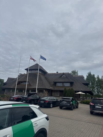 The image shows the Hotel Lilaste in Lilaste, Latvia. The hotel is a three-story building with a thatched roof and multiple windows. In front of the hotel are three flagpoles, with the flags of the European Union and Latvia displayed. Several cars are parked in front of the hotel, including a white van with a green stripe. The sky is overcast. The ground is paved with stone bricks.