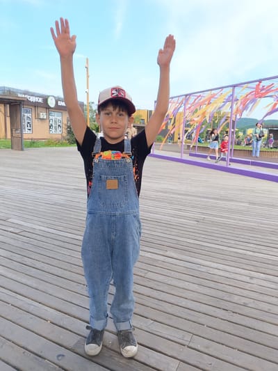 A young boy stands on a wooden deck, raising both arms in the air. He is wearing a black t-shirt, denim overalls, a baseball cap, and sneakers. In the background, there are people, buildings, and a colorful structure with hanging ribbons. This scene is located in the city of Bolshoy Kamen, Russia, on a boardwalk area with recreational facilities.