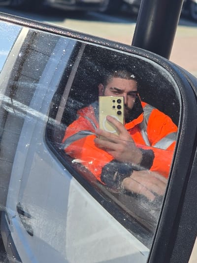 A male individual is reflected in the dirty, scratched side mirror of a white vehicle in Wittenberg, Germany. The subject has dark hair and a beard, and is wearing an orange high-visibility jacket with reflective stripes. He is holding a light-colored smartphone with a multi-lens camera in his right hand, angled towards himself as if taking a self-portrait. His left arm rests within the vehicle's interior, visible through the lower portion of the mirror. The background beyond the vehicle and reflection appears to be an outdoor, brightly lit setting with blurred objects, possibly other parked vehicles.
