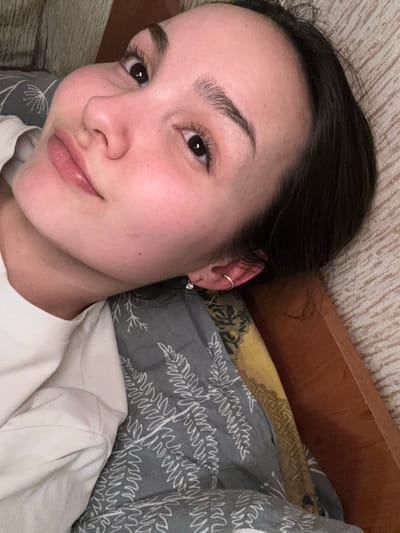 The scene depicts a female individual in an indoor domestic setting, identified as being in Parizhskaya Kommuna, Russia. The individual, appearing to be a young adult with dark hair, fair skin, and dark eyes, is positioned lying down, with her head tilted to the right of the frame, facing and looking directly at the camera. Her facial expression is neutral to slightly smiling. She wears a light-colored (likely white or off-white) t-shirt. Her left ear is visible, featuring a small, clear stud earring in the lobe and a plain gold-colored hoop in the upper helix/cartilage. The individual is resting on a bed or similar furniture. A gray duvet or blanket with a repeating white fern pattern covers a portion of the foreground and midground. To the right, a section of a reddish-brown wooden bed frame is visible, adjacent to a smaller piece of fabric with a yellow and dark patterned design. The background consists of a light-colored, vertically textured wall, possibly beige or cream. Soft, ambient lighting illuminates the scene.