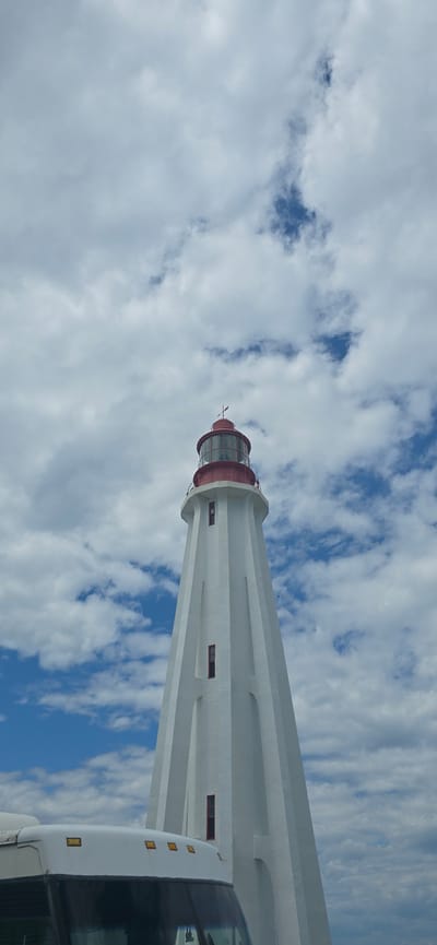 A white lighthouse with a red top stands tall against a cloudy blue sky. The lighthouse is a conical structure with vertical fluting and red rectangular windows. At the very top, a red lantern room is visible, topped with a weather vane. In the foreground, the roof of a white vehicle, possibly a bus, is partially visible, extending from the bottom left corner of the frame. The vehicle has several amber lights on its roof. The lighthouse is located in Rimouski, Canada.