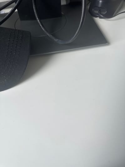 A close-up, top-down view of a white desk surface, located in Thiais, France. In the upper left quadrant, the brim and a portion of the crown of a black fabric baseball cap are visible, exhibiting visible stitching. Centered along the top edge, the dark grey rectangular base of an electronic monitor stand is present. Multiple black electrical cables are routed across and behind this stand; one cable displays white scuff marks. To the upper right, a section of a black electronic device, consistent with a computer speaker featuring a dark grill and a circular button, is partially observed. The desk surface is uniformly lit, casting faint shadows from the objects. No individuals are present, and no actions or interactions are depicted.