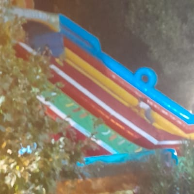 The image shows a close-up, blurred view of a brightly colored inflatable structure, possibly a water slide or bouncy castle, against a dark, out-of-focus background that suggests an outdoor setting at night or in low light. The inflatable features prominent horizontal stripes of blue, yellow, red, and green, with white trim separating some of the colors. A person, appears to be in the upper right portion of the slide, wearing dark clothing. The overall scene suggests a recreational or entertainment venue. The location in Rudəkənar, Azerbaijan is not visually discernible from the provided image.