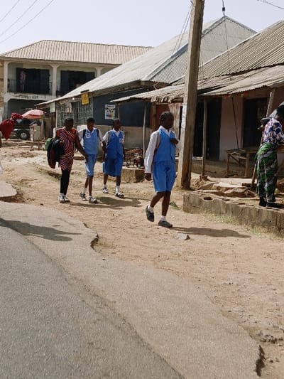 This image captures a daytime street scene in the city of Kungulu School, Zambia. The foreground features a cracked asphalt road on the left, transitioning to an uneven, reddish-brown dirt path on the right.
Four individuals are walking from right to left on the dirt path. The leftmost is an adult female wearing a multi-colored striped top, dark pants, and white shoes, carrying a bundle of fabric over her left shoulder. Adjacent to her are three female students. Two wear blue shorts, a white shirt, and a blue waistcoat, while the third wears a blue skirt, white shirt, and blue waistcoat. All three students wear white socks and athletic shoes, and carry backpacks.
On the far right, a partial view of an adult female wearing a patterned top and green skirt is visible, standing near a wooden structure.
The right side of the street is lined with buildings. The closest is a single-story structure with a grey corrugated metal roof and dark walls, displaying open doorways and wooden fixtures. Behind it, a two-story building with a lighter corrugated roof and light-colored walls is visible. A wooden utility pole with overhead wires stands prominently on the right side of the path. In the far left background, a white vehicle is partially visible next to a red umbrella. The scene is illuminated by strong sunlight, casting visible shadows.