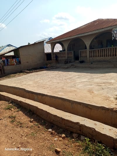 The image shows a dirt road in front of a house in Ilobu, Nigeria, located at latitude 7.835 and longitude 4.471. The house is a single-story structure with a corrugated metal roof and a concrete porch with a railing. There is a single person standing in the doorway of the house. The road is a dirt path with a concrete drainage ditch running along the side. There are some weeds growing in the ditch and on the side of the road. The sky is clear with some white clouds. A sign with "Aramco, Nigeria" is visible in the bottom left corner of the image.