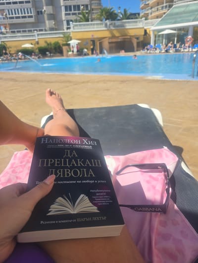 The image, taken from a high-angle perspective, shows a person reclining on a sunbed next to a pool and beach in Peníscola, Spain. The pool is visible in the background, with people swimming and lounging around it. The sandy beach is adjacent to the pool. The person on the sunbed is holding a book titled "Outwitting the Devil" by Napoleon Hill, translated into another language with text in the Cyrillic alphabet, along with a pair of Dolce & Gabbana sunglasses resting on a pink patterned cloth. The person is fair-skinned and wearing an ankle bracelet.