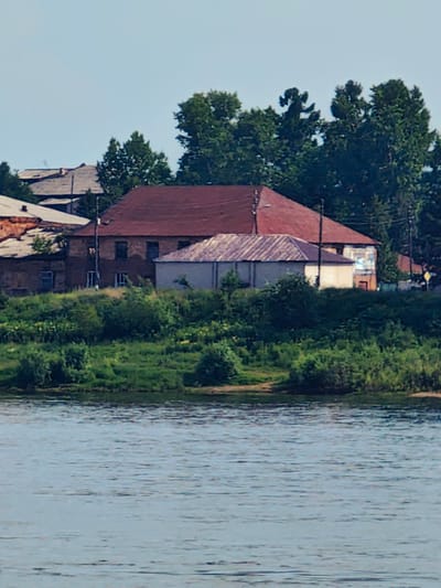 A scene depicts a waterscape in the foreground, with a river occupying the lower portion of the frame. Beyond the river, a lush green embankment rises, densely populated with various trees and shrubs. Perched on this elevated terrain are several buildings, characteristic of a settlement. One prominent structure is a large house with a distinctive red, gabled roof. Adjacent to it is a smaller, lighter-colored building with a corrugated roof. Further to the left, parts of other buildings, including a brick structure with a grey roof, are visible. The sky above is a clear, pale blue. The overall impression is of a riverside community, possibly in the city of Yeniseysk, Russia, characterized by its architecture and abundant natural foliage.