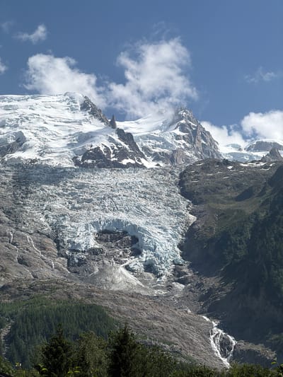 The image depicts a vast glacial landscape at a high altitude, with snow-capped mountains dominating the background under a blue sky with scattered clouds. A prominent glacier flows down the mountainside, its textured surface showing crevasses and ridges of ice. Rocky slopes, partially covered in vegetation, flank the glacier. In the foreground, a dense forest of green trees is visible, with a waterfall cascading down the rocky terrain. The scene suggests a natural, mountainous environment, likely in the vicinity of Chamonix-Mont-Blanc, France, given the characteristic glacial features of the region. There are no people or animals present in the image, and the focus is entirely on the geological and natural elements of the landscape.