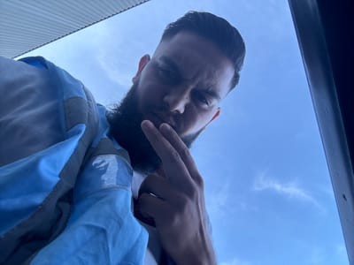 A low-angle, close-up, eye-level shot features a person from the chest up, looking down towards the camera with a neutral expression. The person has dark hair styled with a fade, a full beard, and is wearing a light blue and grey jacket over a white shirt. Their right hand is raised, with two fingers extended, held near their face. The background is a clear blue sky, with a thin, wispy cloud formation visible. A dark, vertical structure is present on the right edge of the frame, and a corrugated metal overhang is visible in the upper left corner. The setting appears to be outdoors, possibly on a street or balcony. There is no information about the location in Boom, Belgium.