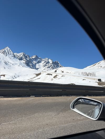 The image, taken from inside a car, shows a view of the snow-covered mountains near Le Monêtier-les-Bains, France. The blue sky is visible through the car window. A snow-covered mountainside is visible next to the road. Part of the road, the roadside barrier, and the car's side mirror are also visible. The image is taken during daylight.