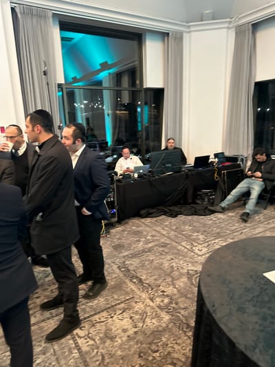 A group of men, likely Jewish, are dressed in dark clothing, some wearing kippahs. They stand in a room with large windows reflecting a turquoise light. In the background, a DJ booth is set up with equipment, and a person is seated on a road case. A large table is partially visible in the corner, covered in a black tablecloth. The room is carpeted with a patterned rug.