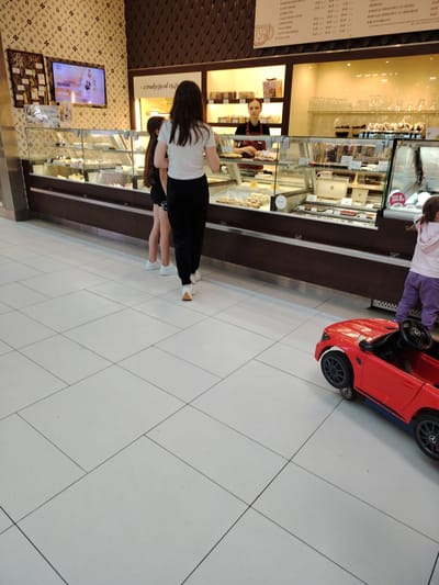 The image depicts the interior of a bakery or cafe with a display case full of pastries. Two young girls, one in a white top and black pants, and another in a black outfit, stand in front of the display. A woman in an apron stands behind the counter, serving customers. In the foreground, a child is sitting in a red toy car. The environment appears to be a well-lit indoor space with tiled flooring and patterned walls. The bakery is located in Wołomin, Poland, as indicated by the presence of Polish text on the display and menu boards.