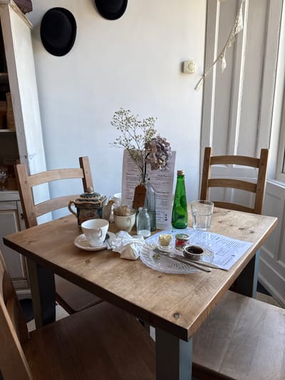 This image presents an indoor scene within a cafe or tearoom located in Ambleside, United Kingdom, specifically focusing on a rectangular wooden table set for, or recently used for, a meal. No individuals are present.

The light brown wooden table supports various items. On the left side, a white porcelain teacup with a floral pattern rests on a matching saucer, containing a silver spoon. Adjacent to it are crumpled white paper napkins. Further back on the left, an ornate ceramic teapot featuring blue, gold, and floral designs is positioned next to a small white creamer and a white bowl filled with sugar sachets. In the center, a clear fluted glass plate holds a used butter knife and a tea strainer, alongside two small jars with remnants of food, possibly jam and cream. A green glass bottle with a cap and an empty clear glass tumbler are located on the right side of the table. Two printed menu sheets are spread out. A tall clear glass bottle contains a dried floral arrangement of baby's breath and a large, muted purple hydrangea bloom.

Three unoccupied wooden chairs with slatted backs are arranged around the table; two are on the left, and one is on the right. The background features white painted walls. On the left, a tall white cabinet with open shelves is partially visible, and two black bowler hats are mounted as wall decor. On the right, a white paneled door or window frame is present, with a small white lace bunting strung across its upper corner. A cream-colored circular thermostat is mounted on the wall above the floral arrangement.