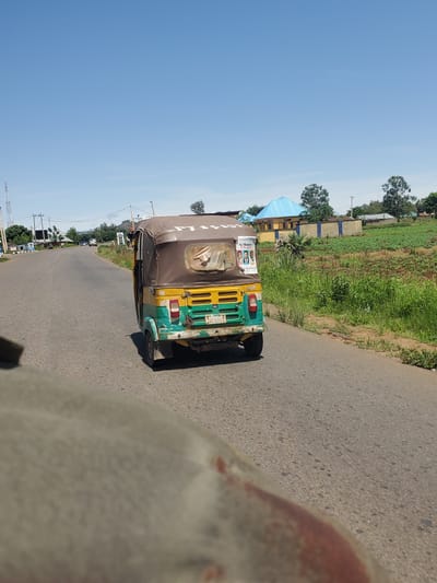 This media file shows a three-wheeled vehicle, commonly known as an auto-rickshaw or "keke," traveling on an asphalt road. The vehicle is predominantly yellow and green, with a brown fabric canopy. Visible on the rear of the auto-rickshaw are advertisements with images of people and text. The road appears to be in a rural or semi-urban setting, with a grassy field and some buildings on the left side of the road. Trees are also present in the background. The sky is clear and blue, suggesting it is daytime and the weather is fair. The camera perspective seems to be from another vehicle moving alongside or slightly behind the auto-rickshaw. The image is oriented vertically, with the sky at the left and the road and auto-rickshaw filling the right side. There are no discernible emotions conveyed, and the scene appears to be a typical day in a Nigerian location, likely Panyam, given the context.