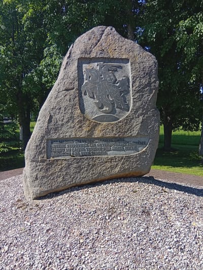 The image shows a monument in Dirvonėnai, Lithuania. The monument is a large, rough-hewn stone with a carved emblem and text. The emblem, located in the upper portion of the stone, appears to be a coat of arms featuring a horse and shield. Below the emblem, the year "2018" is carved. A rectangular plaque with inscribed text is situated below the emblem and year. The monument is surrounded by a bed of small, light-colored pebbles. In the background, lush green trees and grass are visible under a bright sky, suggesting an outdoor setting on a sunny day. The overall scene depicts a historical or commemorative marker in a natural, park-like environment.