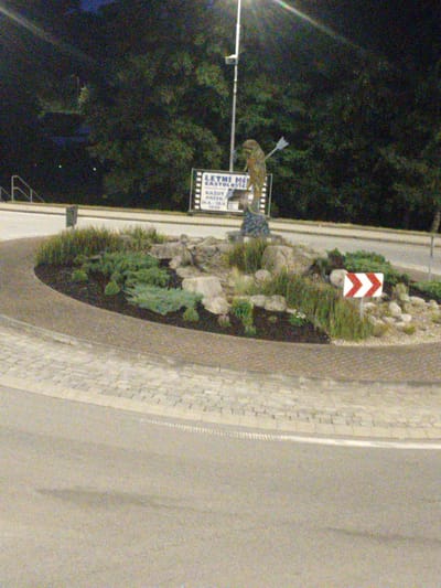 The image shows a roundabout in the city of Častolovice, Czechia. The roundabout has a small landscaped island in the center with rocks and plants. A statue of a person with an arrow is placed on the island, as well as a sign that says "LETNI HIT STOLOTVIC". There is a traffic sign with two red and white chevrons pointing to the right, indicating a sharp turn or obstruction ahead. The roundabout is paved with bricks, and a road runs around it. The background consists of trees and a street light. The image appears to have been taken at night, as the sky is dark and artificial lights are visible.