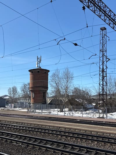 The image depicts a railway environment in Kropachyovo, Russia, under a clear blue sky. In the foreground, multiple sets of parallel railway tracks with dark gravel ballast are visible, alongside a reddish-brown concrete railway platform. A gray metal fence separates the immediate railway area from the midground.
The central subject is a cylindrical, two-story brick water tower, predominantly red-brown. Its upper section features a metal superstructure supporting multiple cellular telecommunication antennas, including panel antennas and several circular dish antennas. Windows are visible in the tower's facade. To the right of the tower, a large lattice steel pylon, part of the railway's overhead electrification system, stands tall, supporting a complex network of overhead power lines and catenary wires with associated insulators.
Bare deciduous trees, indicating a late winter or early spring season, are scattered across the midground. Patches of white snow are visible on the ground, particularly behind the fence. In the background, beyond the trees, several residential or utility buildings are partially visible under the clear blue sky. No individuals, actions, or interactions are present in the scene.