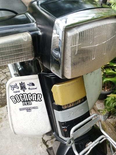 A close-up, slightly elevated view depicts the front section of a black and white Suzuki Deluxe scooter in Salai Har Htan, Myanmar. The vehicle's rectangular headlight, enclosed by a chrome bezel, appears aged with minor dirt and internal striations. To the left, a smaller clear rectangular turn signal is visible. The scooter's body transitions from a glossy black upper section to a matte white lower panel. This white panel is covered with multiple decals, including a prominent black and white sticker that reads "BOENGAR 1931 KAMPUNG PANGERAN TUJUH," surrounded by smaller logos such as "Supra!", a stylized "M" with a cross, "THE NORTH FACE," and "Ashbiel." Below the black bodywork, a gold-colored plastic component with "SUZUKI 33110-38A" embossed on it is positioned above a silver-colored panel bearing "SUZUKI" and "Deluxe" in a cursive font. A silver-toned metal front rack, exhibiting minor rust, is affixed beneath this panel. The immediate foreground shows a light gray concrete or paved surface, while the blurred background on the right contains green foliage and potted plants.
