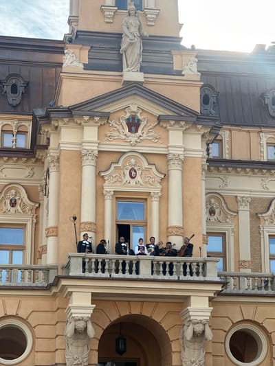 A group of musicians, likely a mariachi band, is performing on a balcony of the historic Sokoł Hall in Nowy Sącz, Poland. The building's facade is a light ochre color with ornate architectural details, including columns, decorative carvings, and statues. The musicians, dressed in traditional mariachi attire, are playing instruments such as guitars and trumpets. One individual is standing near a microphone on a stand, suggesting a performance for an audience below or for recording purposes. The sky above is visible, with hints of sunlight breaking through. The balcony is adorned with a railing, and the overall scene depicts a cultural event or performance taking place on the exterior of the renowned building.