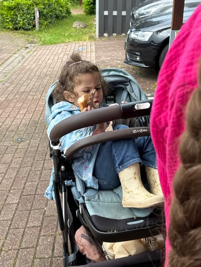 The image captures a young child in a Cybex stroller on a paved walkway in Deinste, Germany. The child, with curly brown hair partially tied up with a small pink clip, is wearing a light blue hooded jacket with a white pattern, blue denim jeans, and light yellow rain boots decorated with a subtle pattern of brown branches and yellow berries. The child holds a piece of light-colored bread or pastry in their right hand, bringing it towards their mouth, and a small, brown, deer-shaped toy in their left hand. The "Cybex" logo is visible on the stroller's brown handlebar. Small green debris is scattered on the stroller's footrest. Beneath the seat, a pink garment with the word "FRIENDS" and a brown paper bag are stored. To the right of the frame, a blurred section of a person is visible, characterized by a vibrant pink textured garment and brown braided hair. The background features a brick-paved path, a strip of green grass, dense green bushes, and a dark fence. Behind the fence, a black sedan is parked or moving slowly, with its front bumper and headlight area partially visible.