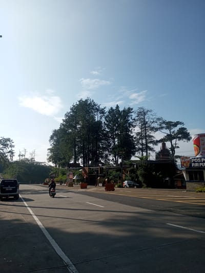 The image shows a road scene in a location that could be Phouviang-Gnai, Laos. The sky is predominantly light blue with some wispy clouds. A road stretches into the distance, with a white line marking the edge of a lane. A black SUV with visible license plate number "F 1897 YP" is driving to the left side of the road, and a person is riding a motorcycle in the same direction.
In the background, there is a building with a sign reading "POS PUR". The building is partly obscured by trees. There are also some trees situated in the middle ground, and a vehicle parked on the side of the road in the distance. There are crosswalks with yellow lines. The light suggests it is either early morning or late afternoon. There are some orange pylons marking the crossing.