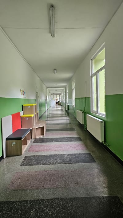 The image depicts a long, straight corridor. The left wall is white and lined with evenly spaced fluorescent tube lights. The ceiling transitions from white to green. A series of rectangular skylights are set into the ceiling. The right wall features vertical bands of varying colors. Several small, open wooden storage units are positioned along the floor. The floor alternates between green and white, with patches of red. The corridor extends into the distance. There is a person at the end of the corridor. The location is Hristo Danovo, Bulgaria.