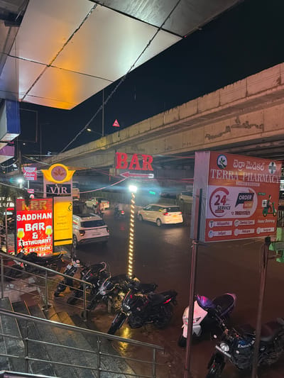 The image shows a street scene at night in Shamshabad, India, under a flyover. Neon signs are prominently displayed, advertising "BAR" in red letters, "Hotel VIR Residency" with a lotus logo, and "SADDHAN BAR & RESTAURANT A.C SECTION" with images of beer glasses. A large banner for "TERRA PHARMACY CHEMIST & DRUGGIST" is also visible, featuring a 24-hour service and home delivery. Several motorcycles and scooters are parked on a wet street, indicating it may have rained. Cars are driving on the road, and a white SUV is parked. The environment appears to be a busy urban area with commercial establishments. The flyover overhead is concrete, and strings of lights are visible along the side of the road.